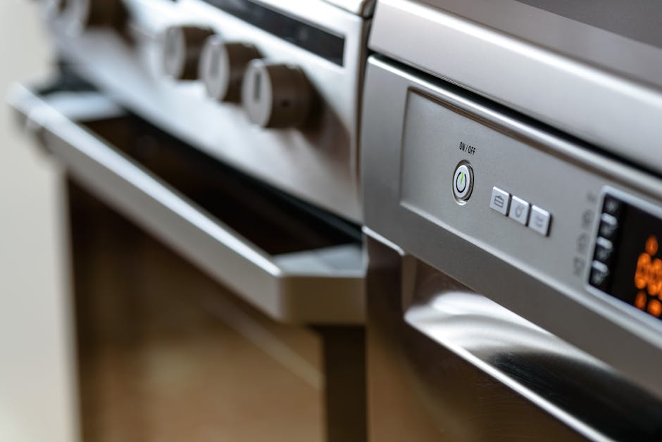Close-up view of a stainless steel oven in a modern kitchen, with a partially open oven door and the control panel visible. The surface appears clean and polished, reflecting light for a shiny finish. In the background, part of a countertop and a nearby oven control knob can be seen. The lighting highlights the smooth, metallic texture of the oven, emphasizing its cleanliness and maintenance. This image illustrates surface cleaning and maintenance aspects related to domestic oven sanitisation, consistent with the services offered by Oven Cleaning Putney, especially in the context of bulky oven parts disposal and deep cleaning procedures.