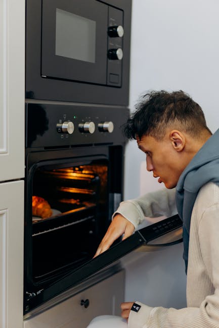 A young man with short dark hair, dressed in a light beige hoodie and cream-colored pants, is kneeling on the light wooden floor of a modern kitchen. He is opening the door of a built-in, stainless steel oven with a digital control panel, set within a white fitted kitchen cabinet. The oven's interior is illuminated, revealing racks and clean cooking surfaces. The kitchen countertop adjacent to the oven is smooth and white, with a clean, minimalist appearance. The room is well-lit with ambient lighting, highlighting the polished surfaces and pristine condition of the appliances, emphasizing the importance of surface cleaning and maintenance as part of domestic cleaning routines. This scene reflects the professional cleaning services offered by Oven Cleaning Putney, specializing in thorough oven care and disposal of bulky oven parts, ensuring hygiene and safety in residential environments.