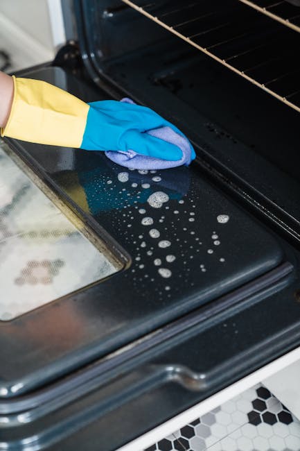 Close-up view of a person wearing a yellow and blue rubber glove using a blue cloth to clean the black stovetop surface of an oven, which appears to be part of a domestic kitchen. The surface shows some soap and water bubbles, indicating recent cleaning or deep surface cleaning for thorough sanitisation. The oven's interior rack is visible, with a reflective metal finish. The countertop materials include a speckled stone surface. The kitchen flooring features black and white hexagonal tiles, adding a modern touch. Bright, natural lighting highlights the shiny, well-maintained appearance of the oven and surrounding surfaces. A natural extension of the detailed cleaning process, this image emphasizes hygienic surface cleaning in a domestic environment, aligning with Oven Cleaning Putney's professional cleaning services.