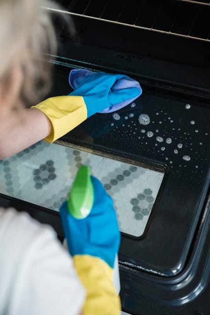 Close-up view of a person wearing a yellow and blue rubber glove using a blue cloth to clean the black stovetop surface of an oven, which appears to be part of a domestic kitchen. The surface shows some soap and water bubbles, indicating recent cleaning or deep surface cleaning for thorough sanitisation. The oven's interior rack is visible, with a reflective metal finish. The countertop materials include a speckled stone surface. The kitchen flooring features black and white hexagonal tiles, adding a modern touch. Bright, natural lighting highlights the shiny, well-maintained appearance of the oven and surrounding surfaces. A natural extension of the detailed cleaning process, this image emphasizes hygienic surface cleaning in a domestic environment, aligning with Oven Cleaning Putney's professional cleaning services.