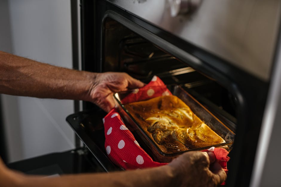 A person's hand is placing a glass baking dish with roasted chicken into an electric oven for surface cleaning and deep cleaning. The oven interior appears clean and well-maintained, with a black enamel surface and oven racks. The baking dish rests on a red and white polka-dot oven mitt, indicating careful handling. The surrounding kitchen area features neutral-colored tiles and countertop, with natural lighting illuminating the scene, emphasizing hygiene and maintenance associated with domestic cleaning services like those offered by Oven Cleaning Putney. This image reflects thorough oven sanitisation and surface cleaning, suitable for highlighting emergency oven deodorising and same-day oven cleaning options in Putney.