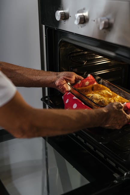 A young man with short dark hair, wearing a beige hoodie with a grey sweatshirt draped over his shoulders, is kneeling on a light wooden floor while open-in the oven in a modern kitchen. The oven's stainless steel door is open, revealing a clean interior with a warm glow from the heating element. To the left, there is a black microwave above a built-in stainless steel oven, both set into white cabinetry. The kitchen wall behind him is plain white, and there is bright, even lighting that highlights the surfaces' cleanliness. The scene depicts surface cleaning and maintenance of kitchen appliances, consistent with services like those offered by Oven Cleaning Putney for deep cleaning and sanitisation in residential settings.