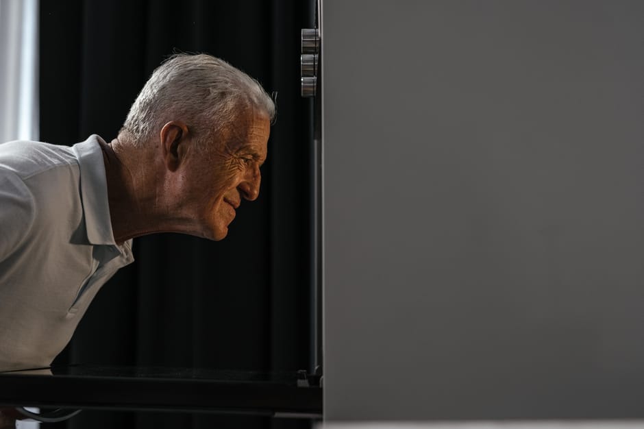 A mature man inspecting the interior of a kitchen oven, which is built into a grey kitchen unit. The oven door is open, revealing a clean, shiny black surface, possibly stainless steel or ceramic, with a reflective finish. The man is leaning in closely, with a focused expression, wearing a light-colored shirt. The surrounding kitchen area has a contemporary design with dark cabinetry and subtle lighting, emphasizing the cleanliness and maintenance of the oven. This image illustrates surface cleaning and deep cleaning processes aimed at tackling stubborn stains, aligning with Oven Cleaning Putney's services in surface sanitisation and domestic cleaning, specifically addressing persistent oven stains as discussed in the 'Grease Rebuilds' article on SW15, PUTNEY.