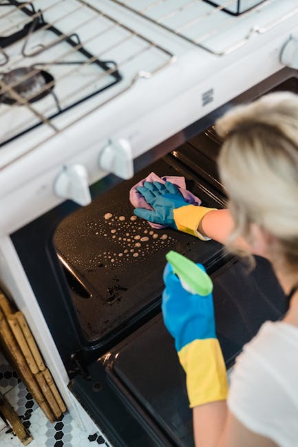 A person wearing blue and yellow rubber gloves is cleaning the interior of a black oven with a pink cloth. The oven door is open, revealing a dark oven cavity with visible grease and stains on the oven floor. The stovetop above the oven features a white surface with black burners and control knobs. The kitchen floor has black and white tile patterns, and part of a wooden cabinet can be seen on the left side. The scene is well-lit, emphasizing the cleaning process, which aligns with deep surface cleaning and sanitisation practices, as offered by Oven Cleaning Putney, to address stubborn oven stains in SW15, Putney.