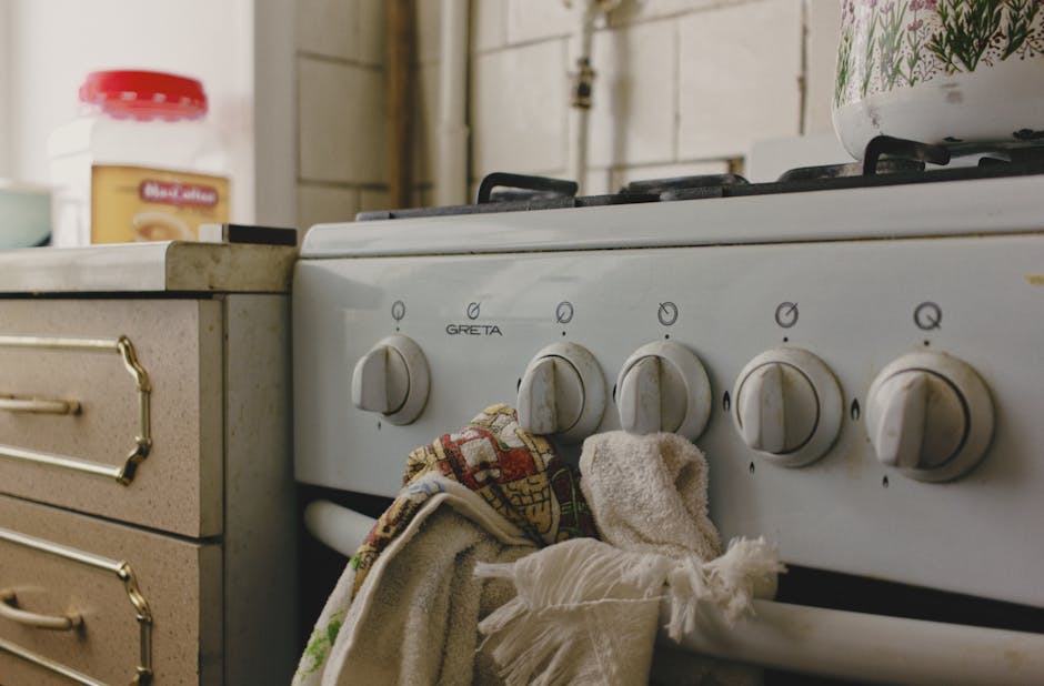 Close-up image of a white gas stove with four round control knobs, situated in a domestic kitchen. The stove surface appears clean with a slight sheen indicating recent sanitisation. A partially folded, patterned dish towel hangs from the oven handle, and in the background, there is a beige kitchen counter with a jar of peanut butter. The counter surface is smooth and free of dust or debris, while the surrounding area shows natural lighting. The kitchen wall behind the stove features white tiles with visible grout lines. The overall scene reflects surface cleaning and maintenance practices, consistent with professional domestic cleaning services such as those provided by Oven Cleaning Putney, in line with their focus on hygiene and deep cleaning for a well-maintained kitchen environment.