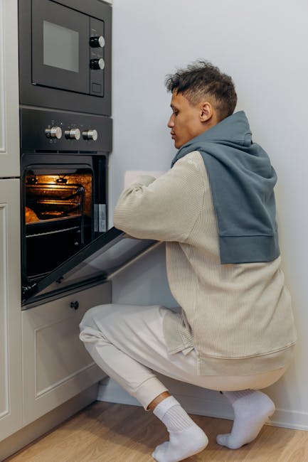 A young man with short dark hair, wearing a light beige hoodie with a grey sweatshirt draped over his shoulders, is crouching in front of a built-in electric oven with a stainless steel finish, located beneath a black microwave. The oven's glass door is open, revealing a clean, illuminated interior with a wire rack. The kitchen features light-colored cabinetry and wooden flooring, and the lighting is bright and even, highlighting the stainless steel surfaces. The man appears to be inspecting or preparing to clean the oven, emphasizing thorough surface cleaning and maintenance, which aligns with the expertise of Oven Cleaning Putney in domestic cleaning services for surface sanitisation and deep cleaning.