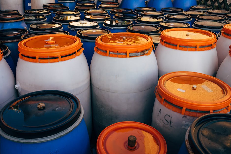 An outdoor storage area containing various plastic containers and cans used for chemical disposal, with colors including blue, yellow, orange, and black. The containers appear to be made of durable pl