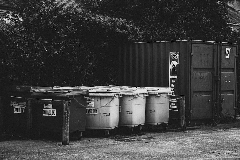 A row of black and white waste bins and recycling containers situated outdoors on a paved surface, adjacent to a large metal storage container. The bins include a mix of paper and general waste, with labels indicating their contents and designated disposal types. The waste bins are made of plastic, with some featuring attached lids, and are positioned in front of dense foliage and trees in the background. The scene is evenly lit, highlighting the clean surfaces of the bins and the surrounding area. The setting appears to be part of a residential or community waste disposal point, reflecting proper waste segregation and collection practices in accordance with local regulations, as discussed by Oven Cleaning Putney in the context of Wandsworth Council rules on oven waste disposal in Putney.