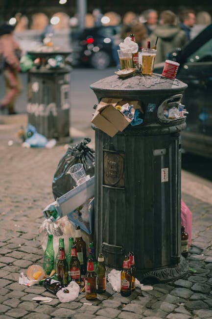 An overflowing black public trash bin made of metal with a domed lid featuring openings for waste, located on a cobblestone street in Wandsworth. The bin is surrounded by scattered litter, including glass bottles, paper cups, food containers, and miscellaneous debris. On top of the lid, several used paper cups with plastic lids, a crumpled paper napkin, and a small cardboard box are visible. To the side of the bin, a trash bag is seen filled with additional waste. In the background, blurred pedestrians and parked vehicles suggest an urban setting, with soft natural daylight illuminating the scene. This image emphasizes the importance of proper waste management and the need for regular cleaning and sanitisation to maintain hygiene in public spaces, aligning with the local regulations outlined by Wandsworth Council on waste disposal, as discussed on the ovencleaningputney.com page about oven waste disposal rules in Putney.